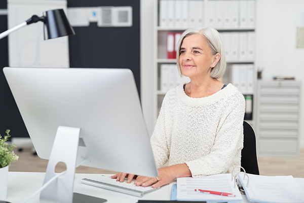 Older Woman Sitting At Desk In Front Of Computer